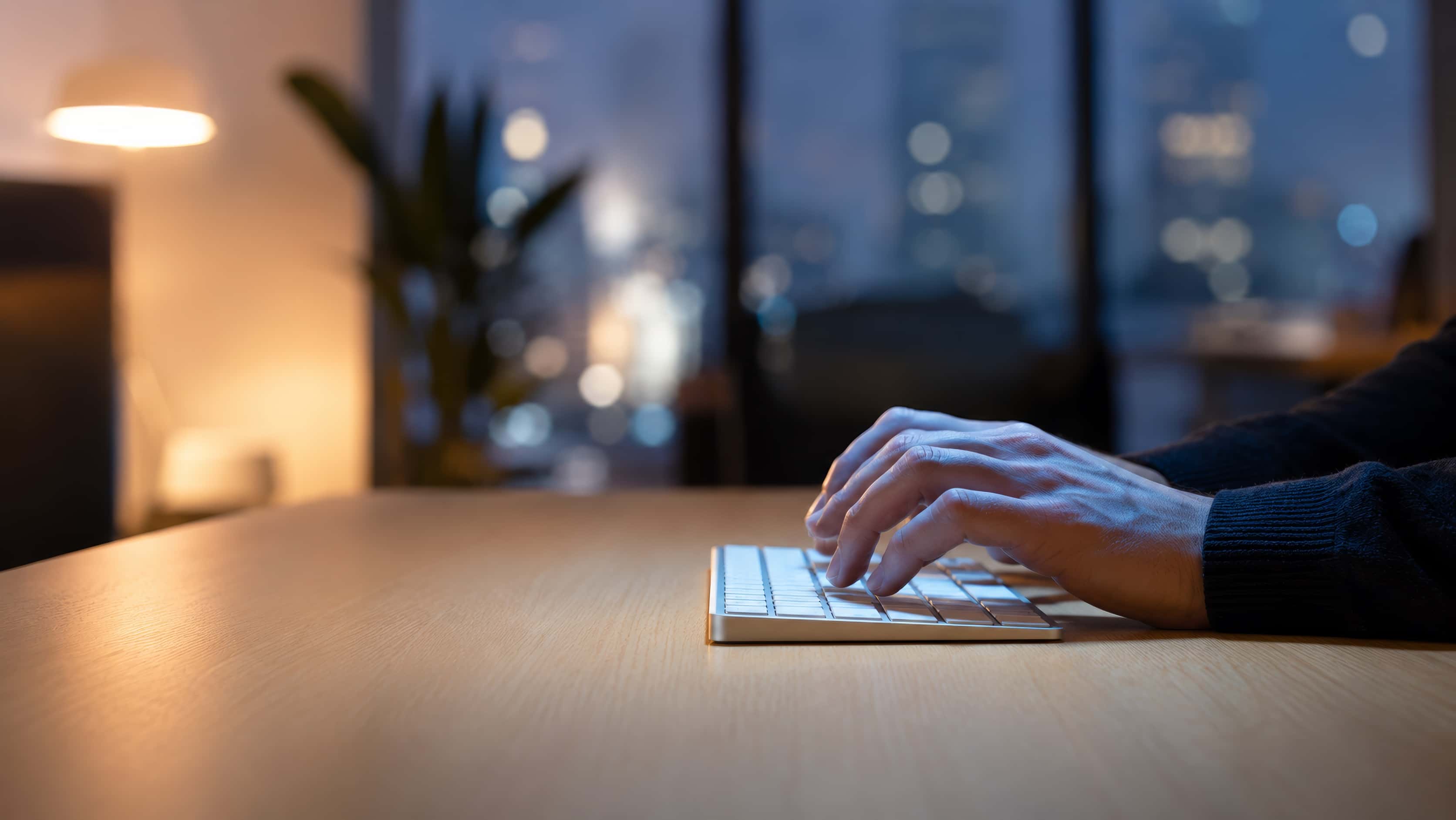 Hands on a low-profile keyboard, in profile, on a clean minimal desk — mixed light of a warm lamp and a cool off-frame screen.